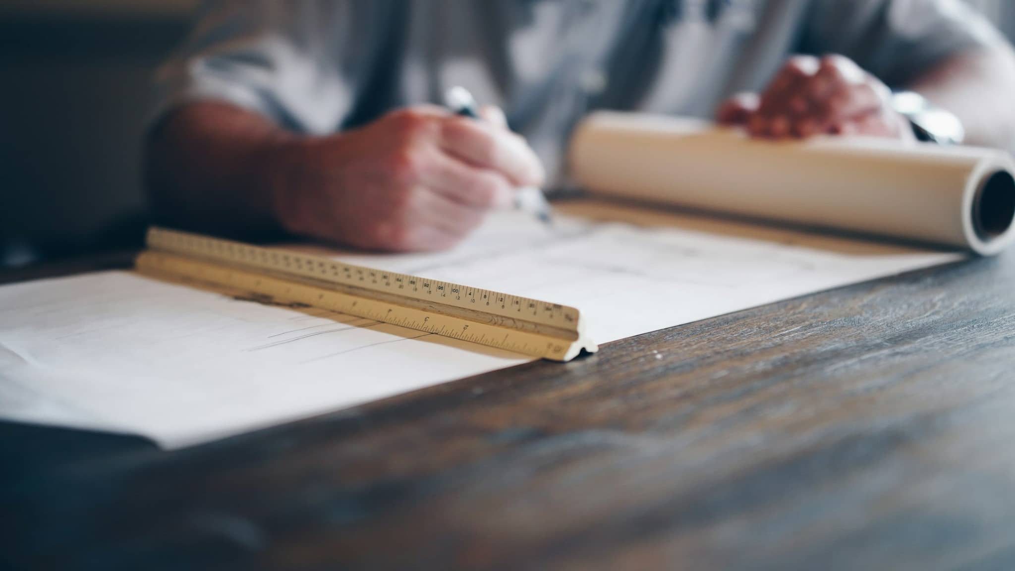 Hands drafting on a roll of architectural drawings at a wood desk